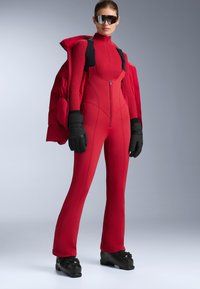Piros síoverall magas nyakkal, elöl cipzárral és testhezálló szabással. Fekete kesztyűvel és síbakancsokkal viselve, vastag piros dzseki réteggel kiegészítve.