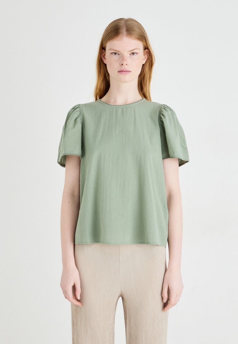 Bluzka s kratkimi rokavi, svetlo zelena, z napihnjenimi rokavi in okroglim izrezom. Narejena iz mehkega blaga z rahlo teksturirano površino.