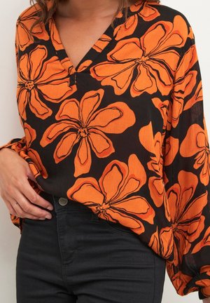 Black V-neck blouse with large orange floral print worn with black pants, hand resting on hip, torso visible.