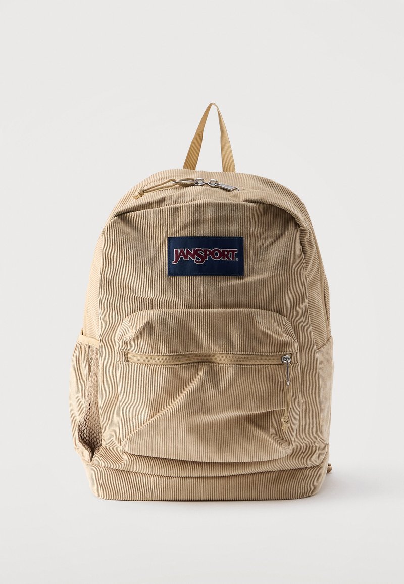 Μπεζ σακίδιο πλάτης Jansport από κορντόρι με εμπρόσθια τσέπη με φερμουάρ, δίχτυ πλαϊνή τσέπη και χειρολαβή στην κορυφή σε λευκό φόντο.