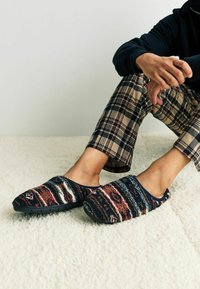 Vlněné pantofle s texturovaným, vícolor vzorem v odstínech červené, modré a bílé; černá gumová podrážka; nošeny s kostkovanými kalhotami na měkkém povrchu.
