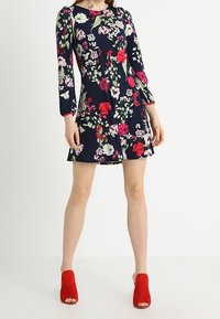 Vaatteet kukkakoristeisella kankaalla, pitkät hihat, tummansininen tausta, monivärisiä kukkia, kapea vartalo, liehuva hame, yhdistettynä punaisiin avonaisiin kengänkorkoihin.