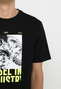 Black cotton t-shirt featuring a graphic of a motorcycle tire and the phrase "MODEL IN INDUSTRY" in bright yellow text.