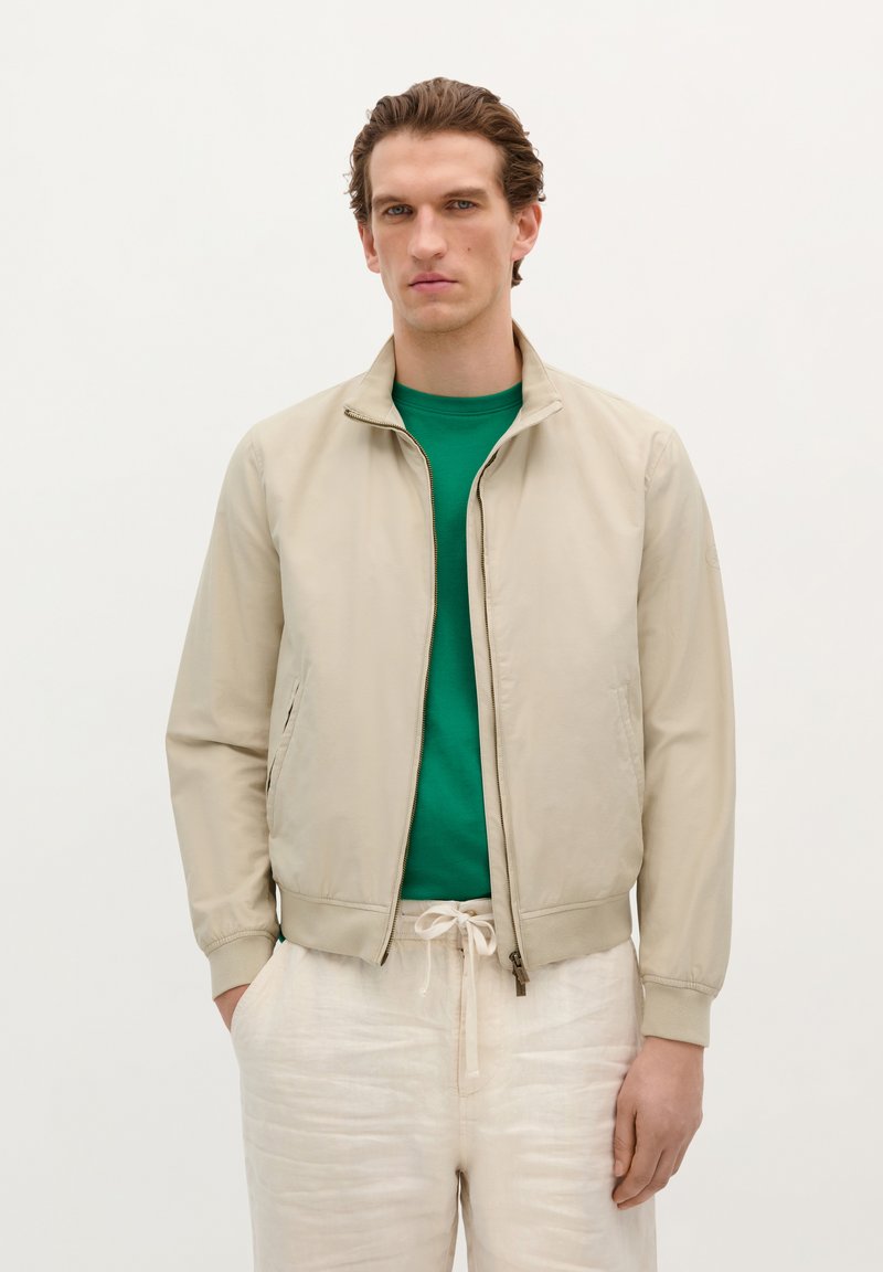 Chaqueta bomber beige con cierre frontal, puños de punto y bolsillos laterales, llevada sobre una camiseta verde. También presenta pantalones sueltos de color claro.