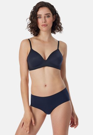 Vrouw met krullend haar die een bijpassende navy blauwe bh en hipsteronderbroek set draagt, staand tegen een effen lichte achtergrond.