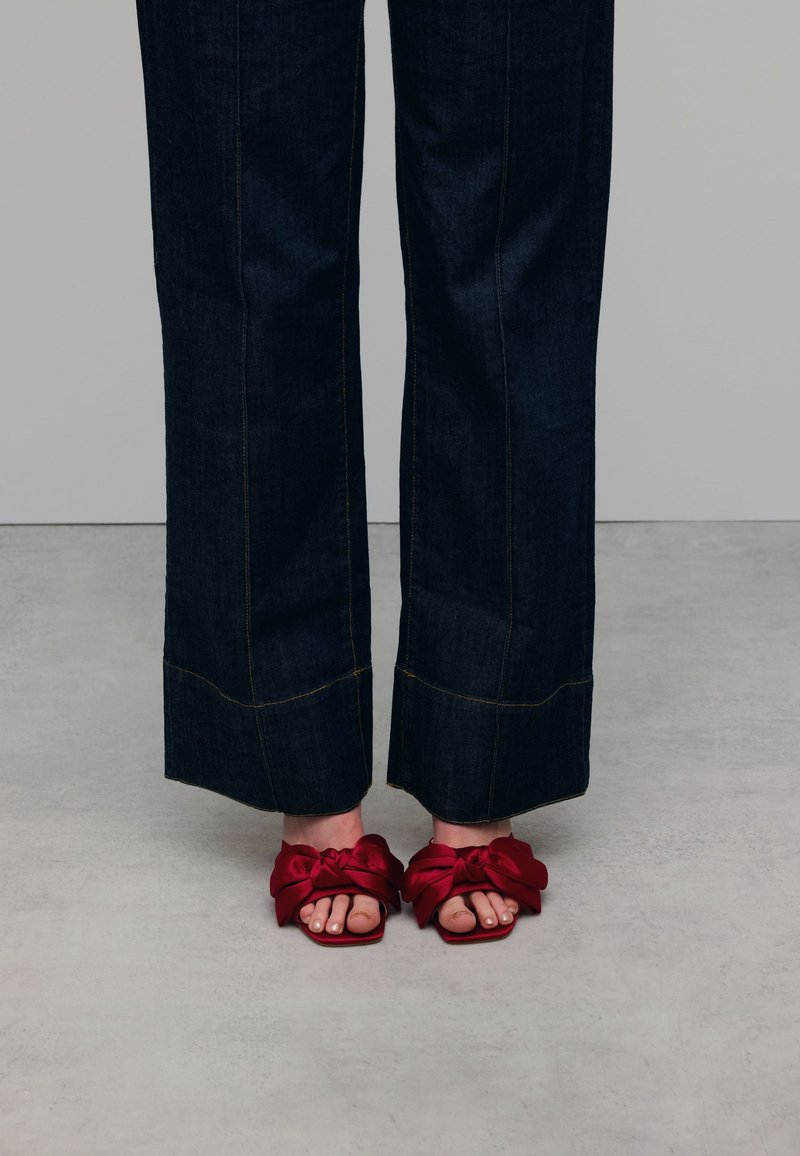 Rode satijnen slidesandalen met oversized strikken aan de voorkant, gecombineerd met donkere denim wijde pantalon met contrasterende stikseldetails.
