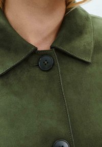 Veste en daim vert olive dotée d'un col pointu, de deux boutons noirs et de surpiqûres contrastantes le long des bords. Texture douce sur toute la surface.