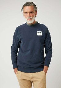 Hombre mayor con barba gris que lleva un sudadera azul marino con el texto "Dream Big Altonadock" y pantalones beige, de pie con las manos en los bolsillos.