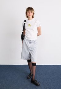 White t-shirt with green graphic design, paired with light denim shorts and black shoes over black knee-high stockings. Dark crossbody bag.