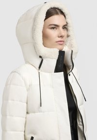 Puffig vit jacka med en stor, fluffig huva klädd i krämfärgad fleece. Har en frontdragkedja, svarta detaljer och ett quiltat mönster.