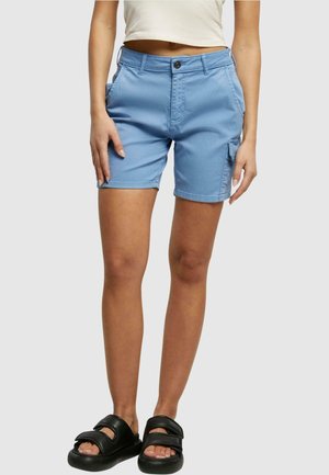Femme portant un short bleu clair au genou avec une poche latérale, un top court blanc et des sandales noires à bout ouvert, debout devant un fond uni.