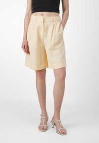Pantaloni in lino giallo chiaro con vestibilità rilassata, fronte plissettato e tasche laterali, abbinati a sandali bianchi con tacco.