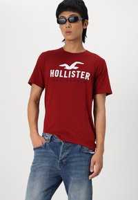 Crvena pamučna majica s bijelim logotipom "HOLLISTER CALIFORNIA", u kombinaciji s plavim trapericama. Model nosi crne sunčane naočale.