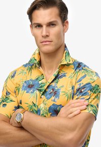 Camisa de manga corta, amarilla, con botones y un patrón floral azul con hojas verdes. El modelo lleva un reloj dorado con una correa de cuero color beige.