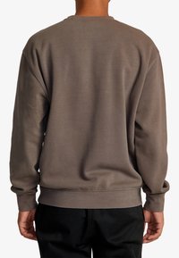 Sweat-shirt marron avec un col rond, des manches longues et des poignets côtelés. Texture douce, design simple et coupe décontractée. Pantalon noir visible.