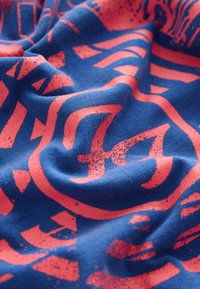 Modro bombažno blago z gladko teksturo ima velike, izrazite rožnate grafične odtise, vključno s črkami in oblikami v obrabljenem slogu.