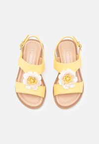 Sandalias amarillas con dos correas anchas, acentos florales en blanco y beige, y una correa en el tobillo con hebilla. Textura suave con detalles de costura.