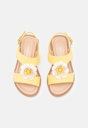 Gelbe Sandalen mit zwei breiten Riemen, floralen Akzenten in Weiß und Beige sowie einem geschnallten Knöchelriemen. Glatte Textur mit genähten Details.