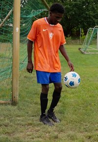 Oranje Nike shirt met witte accenten, blauwe shorts, zwarte sokken, die een witte voetbals bal met blauwe en zwarte markeringen jongleert op een grasveld.