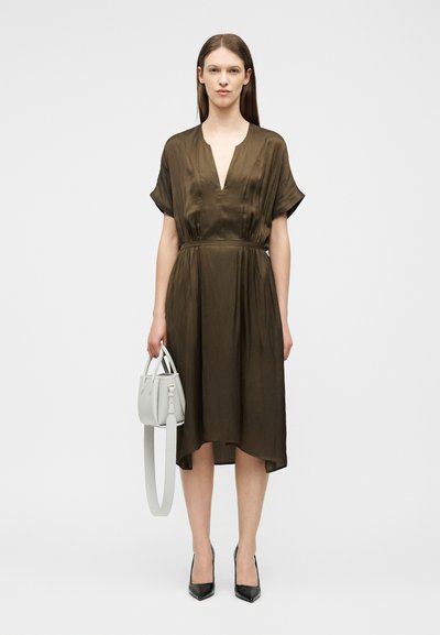 Γυναίκα που φοράει φόρεμα μήκους μέχρι το γόνατο σε ελιά πράσινο χρώμα με κοντά μανίκια, κρατώντας μια λευκή τσάντα και φοράει μαύρες μυτερές ψηλοτάκουνες γόβες.