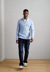 Light blue zip-up sweater, ribbed texture, worn over a beige shirt, paired with dark denim jeans and white sneakers. Simple design, casual fit.