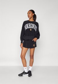 Must musta NikeCourt sweatshirt valge kiri, sobivad mustad lühikesed püksid ja mustad spordijalatsid. Omab pehmet tekstuuri ja logot.