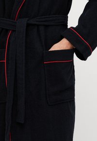 Svart frottébadekåpe med rød piping, belte og lommer; har en myk tekstur og løs passform, designet for komfortabelt bruk.