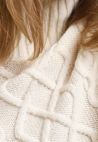Cream-colored knitted sweater featuring a textured, cable knit design with raised patterns. Soft, chunky knit material.