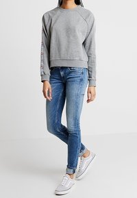 Felpa grigia con maniche raglan e dettaglio del logo, abbinata a jeans fitted blu chiaro e sneakers bianche. Outfit informale e confortevole.