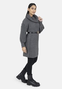 Grå strikket sweaterkjole med høj hals, bælte i taljen med et sort bælte med en gylden hjerteformet spænde, parret med sorte leggings og støvler.