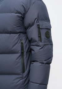 Tengerészkék steppelt kabát, cipzáras zsebekkel az ujjon és hatszögletű logóval a felkaron.