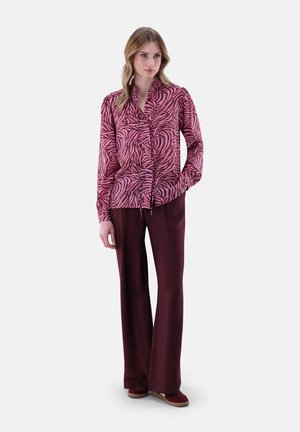 Langärmlige Bluse in Pink mit schwarzem Zebramuster, kombiniert mit bordeauxfarbenen Schlaghosen. Die Bluse hat einen Stehkragen und Knöpfe.