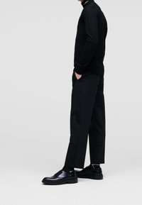 Černý pletený svetr s vysokým límcem, doplněný černými širokými kalhotami s jemným bočním pruhem. Lesklé černé boty doplňují celkový vzhled.