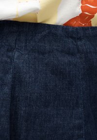 Σκούρο μπλε τζιν ύφασμα κάτω από ένα ανοιχτόχρωμο πουκάμισο με κόκκινο-πορτοκαλί λουλουδένιο μοτίβο, που εμφανίζει λεπτομέρειες υφής και ραφών.