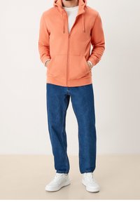 Korallenroter Hoodie mit Reißverschluss und Kordelzug, zwei Fronttaschen; kombiniert mit lockeren blauen Jeans und weißen Turnschuhen; weicher Stoff, lässiges Design.