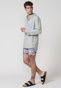 Lichtgrijze overhemd met knopen en een borstzak, bloemen zwemshorts in pastelkleuren, en zwarte sandalen met een kurken zool.