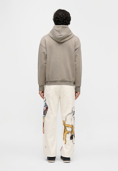 Μπεζ φούτερ με κουκούλα και άνετη εφαρμογή, συνοδευόμενο από εκρού παντελόνι με πολύχρωμες εικονογραφήσεις και στα δύο πόδια. Ορατά μαύρα παπούτσια.