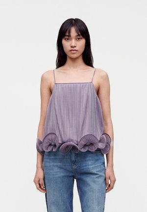 Jeune femme aux longs cheveux foncés porte un haut sans manches plissé couleur lavande avec un ourlet volanté et un jean bleu sur fond blanc uni.