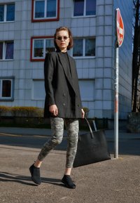 Blazer noir à double boutonnage, leggings camouflage gris, et chaussures noires à enfiler. Sac cabas noir avec texte embossé. Cadre extérieur lumineux.