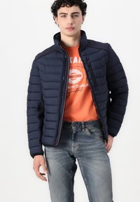 Jachetă puffer navy cu cusături orizontale, guler înalt și buzunare cu fermoar, asortată cu un tricou portocaliu și blugi deschiși la culoare.