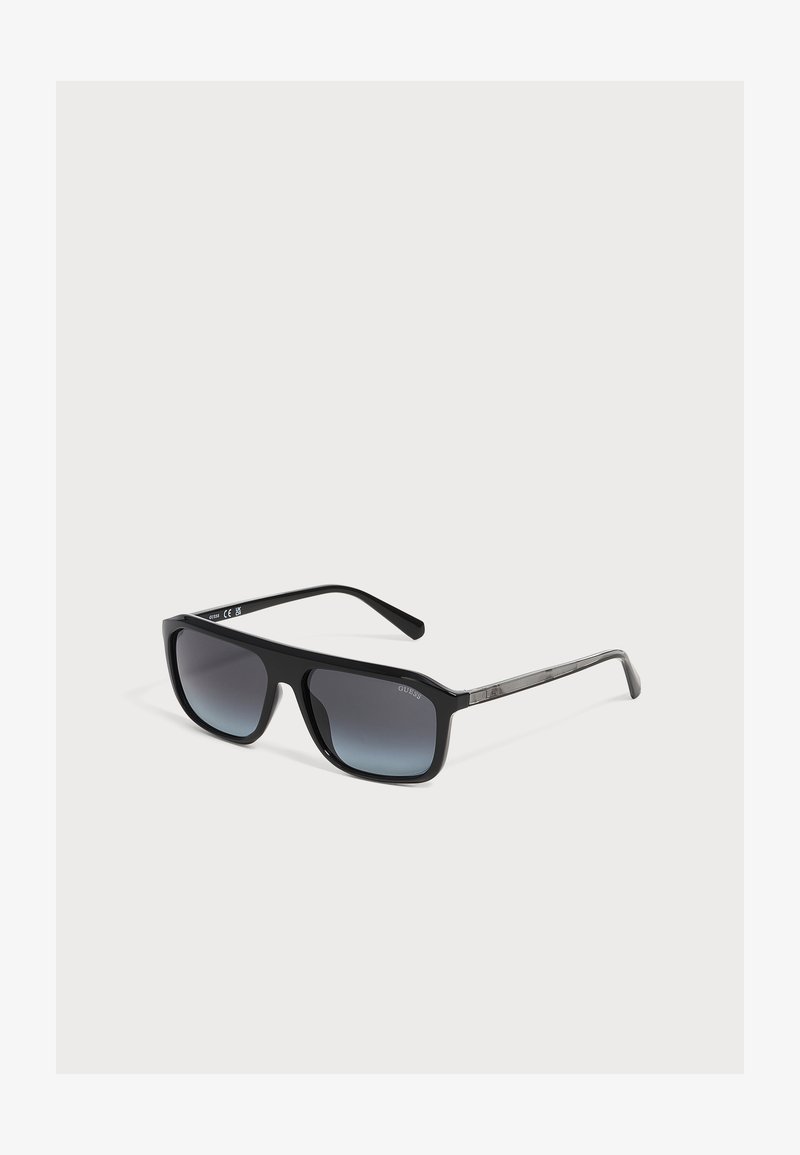 Lunettes de soleil rectangulaires noires avec des verres dégradés foncés et des branches fines, posées sur un fond gris clair.