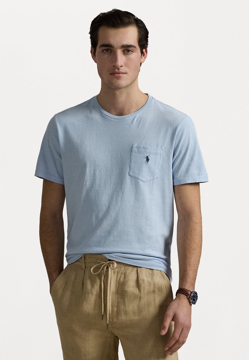 Light blue cotton t-shirt with a round neck, short sleeves, and a chest pocket featuring a small logo. Paired with tan linen drawstring pants.