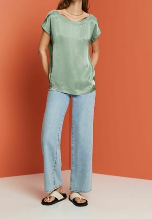 Vaaleanvihreä satiinitoppi, jossa on lyhyet hihat ja leveä pääntie, yhdistettynä vaaleansinisiin leveälahkeisiin farkkuihin ja mustiin sandaaleihin, joissa on valkoiset remmit.