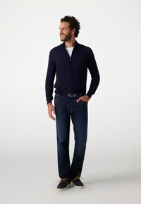 Maglione navy con zip realizzato in tessuto morbido, abbinato a jeans in denim scuro e scarpe navy con lacci marroni. Design semplice e contemporaneo.