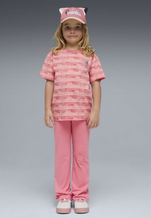 Młoda dziewczynka w różowej czapce z uszami kota, różowej koszulce z wzorem, dopasowanych spodniach z motywem kota oraz białoróżowych trampkach, stojąca na jednolitym szarym tle.