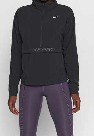 Person trägt ein schwarzes Nike Pro Langarm-Quarter-Zip-Shirt und lila Leggings mit Seitennähten, steht vor einfarbigem Hintergrund.