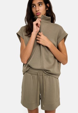 Młoda kobieta o długich ciemnych włosach zapinająca suwaki w bezrękawniku w oliwkowym kolorze, do którego dobrane są krótkie spodenki, na jednolitym tle.