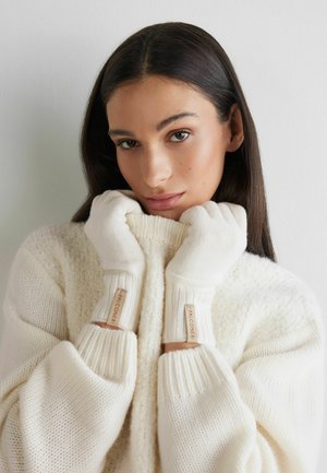Witte handschoenen van een wolmix met geribbelde manchetten en leren labels. Het model draagt een chunky gebreide crèmekleurige trui, met haar handen dicht bij haar gezicht.