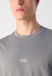 Grå T-skjorte laget av myk stoff, med rund hals og en hvit "HUGO"-logo på brystet, glatt tekstur og tettsittende design.