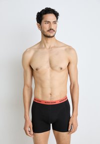 Fekete boxeralsó piros gumírozott derékkal, amelyen a "BJÖRN BORG" logó található. Az anyaga sima, és a szabása testhezálló. A modell álló helyzetben van.